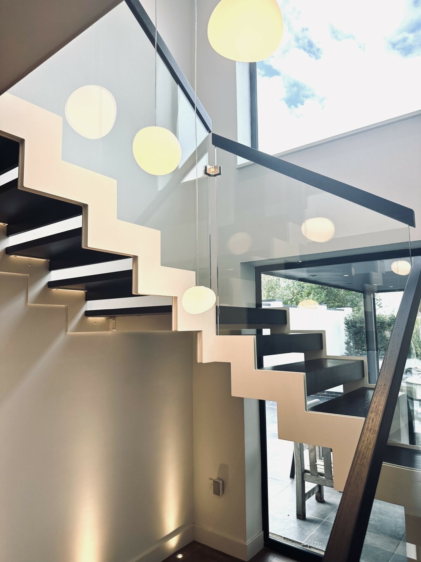 An interior view of a zigzag contemporary staircase featuring stained oak steps, a glass balustrade, and round pendant lights, with a view of the sky through large windows.